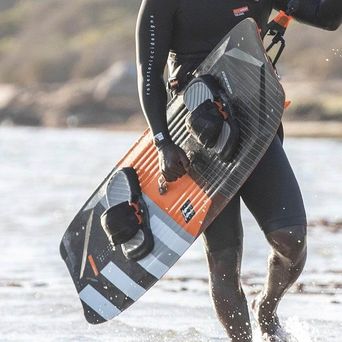 Surfer mit Bord in den Händen am Strand, bereit für das Kitesurfen in der Wasserlandschaft.
