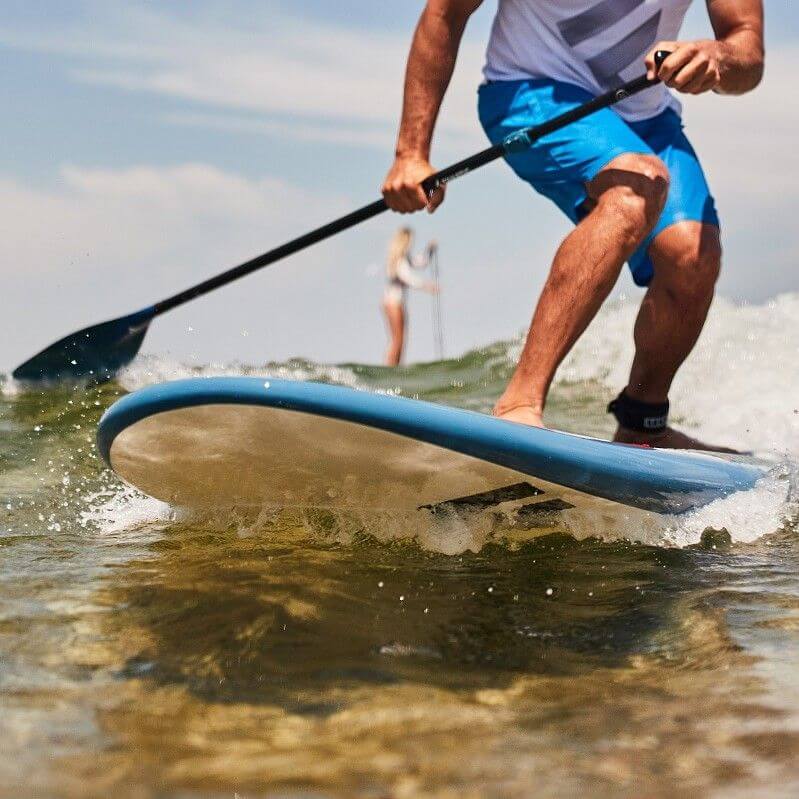 Mann beim Stand-Up-Paddling auf einem blauen Board in sanften Wellen, im Hintergrund eine andere Paddlerin.