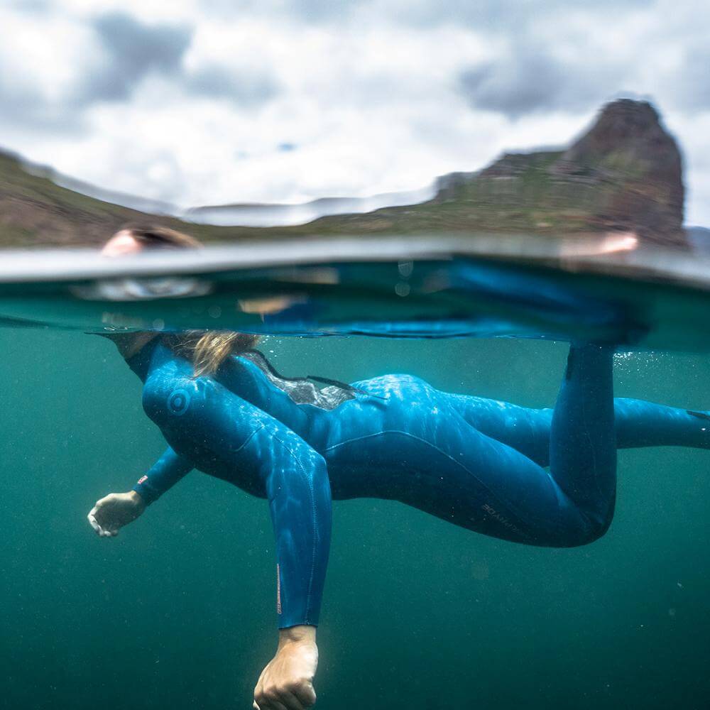 Eine Person in einem blauen Neoprenanzug, die unter Wasser schwimmt, umgeben von klarem Wasser und Bergen.