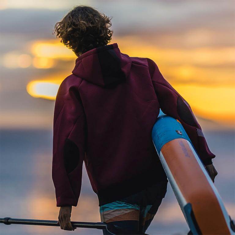 Junger Mann in Kapuzenpullover, der ein Stand-Up-Paddle-Board bei Sonnenuntergang trägt.
