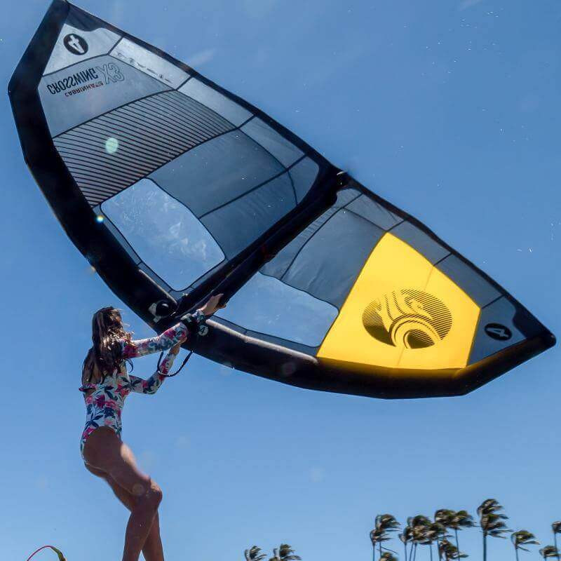 Frau mit einem großen aufblasbaren Wing beim Wassersport unter klarem Himmel, umgeben von Palmen.