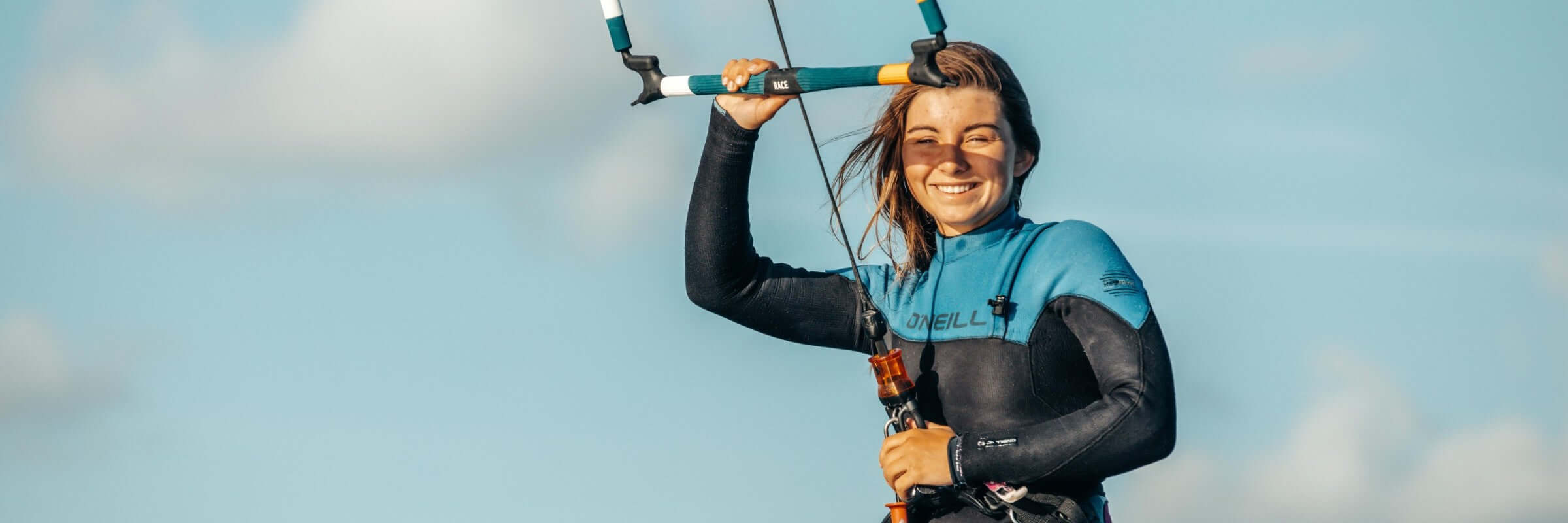 Lächelnde Surferin in einem blauen Neoprenanzug hält das Kiteboard in der Hand vor einem klaren Himmel.