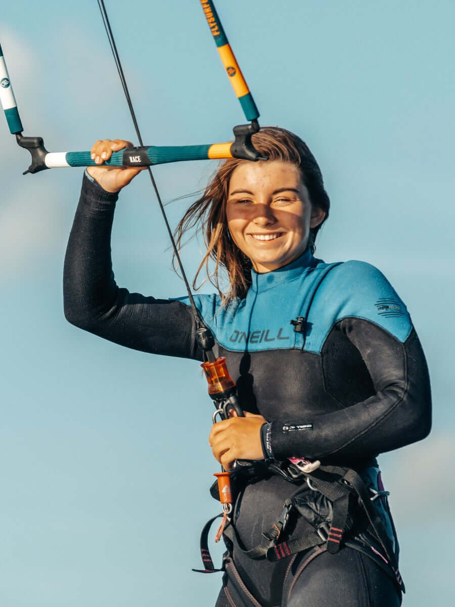 Junge Frau im Neoprenanzug hält Kite-Bars am Strand, strahlend und bereit für das Kitesurfen.