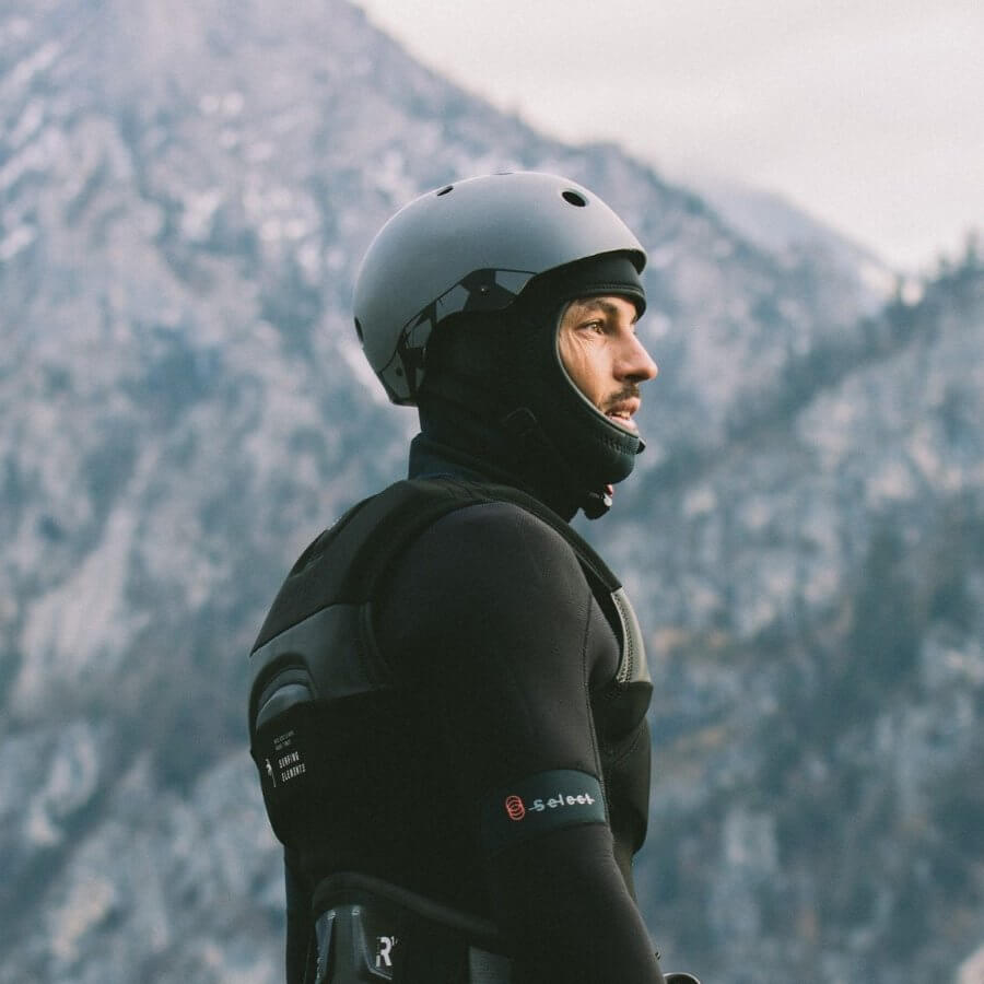 Surfer mit Helm, Prallschutzweste, Trapez und Neoprenanzug, bereit für Wassersport in den Bergen, ideal für einen Online Surfshop.