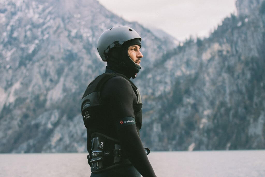 Mann in einem Neoprenanzug und Helm, betrachtet die Landschaft am See mit Bergen im Hintergrund.