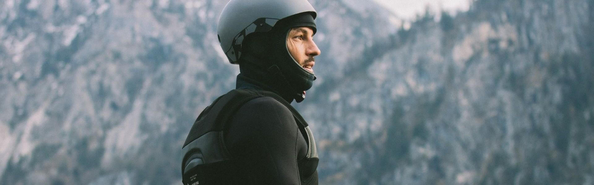 Mann in Neoprenanzug und Helm für Wassersport, bereit zum Surfen in bergiger Umgebung.