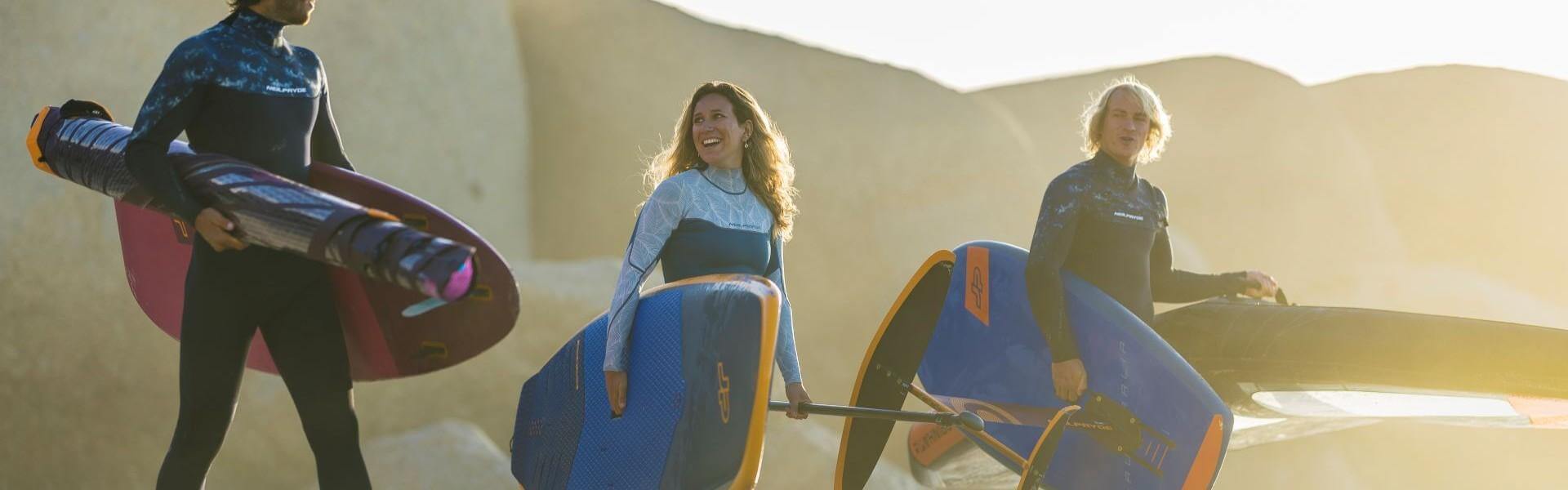 Drei Windsurfer in Neoprenanzügen halten ihre Surfbretter am Strand bei Sonnenuntergang.