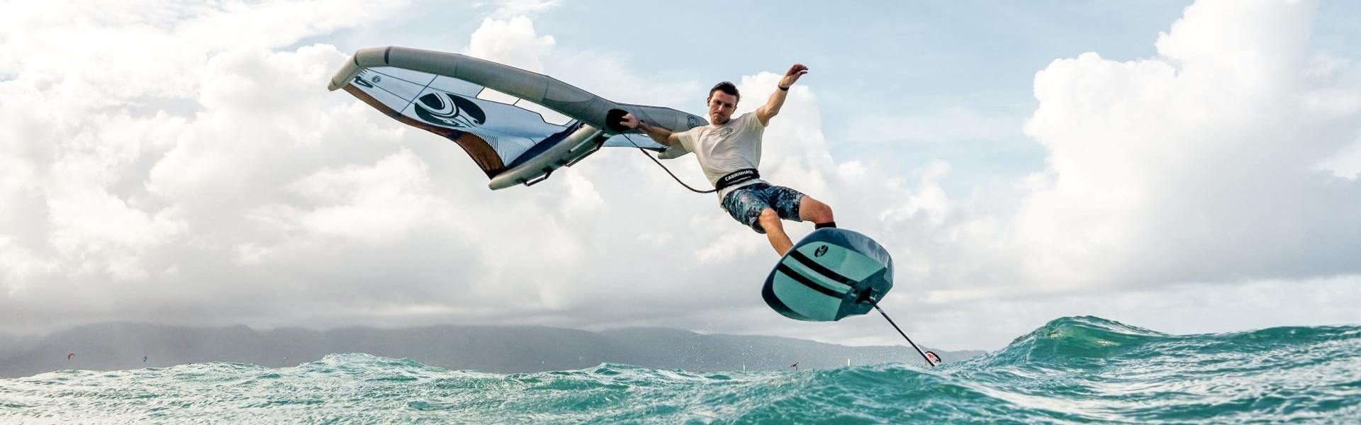 Surfer performing a dynamic trick on a hydrofoil board with a wing in ocean waves under a cloudy sky.