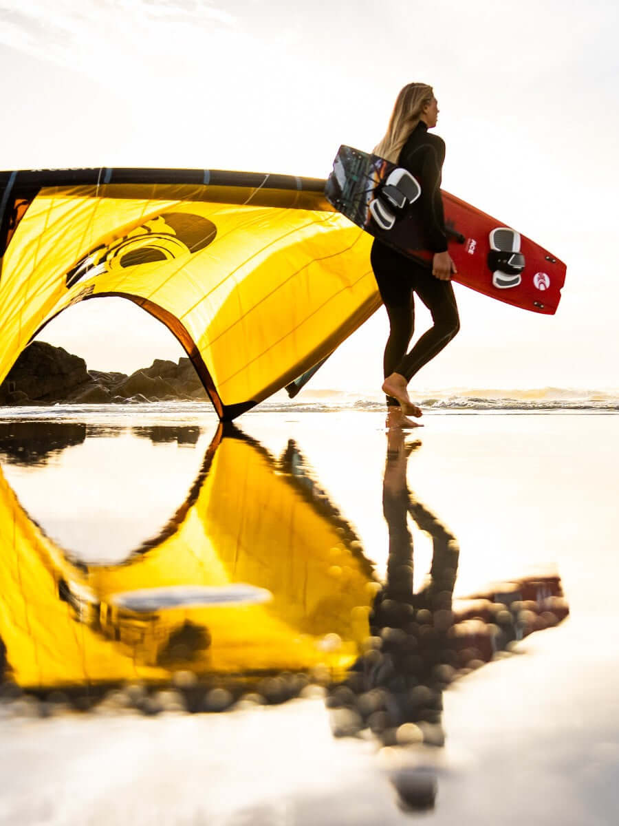 Surferin mit Kite und Board am Strand bei Sonnenuntergang, Spiegelung im Wasser, sportliche Dynamik.
