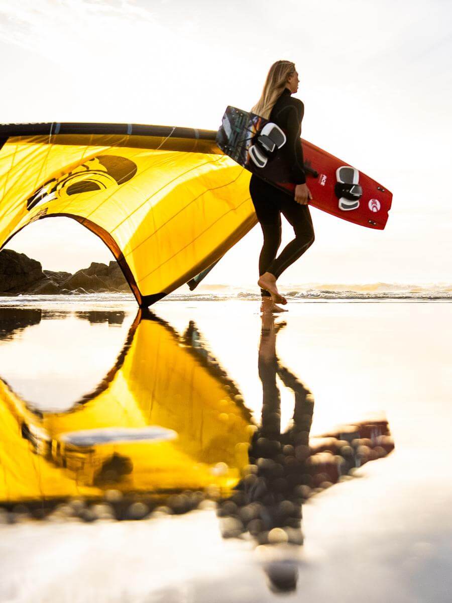 Mädchen geht am Strand mit einem gelben Kite und einem Surfbrett entlang, reflektiert im Wasser.