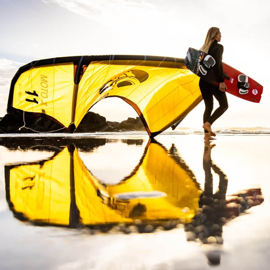 Mädchen geht am Strand mit einem gelben Kite und einem Surfbrett entlang, reflektiert im Wasser.