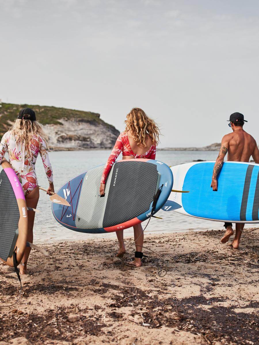 Drei Surfer mit Paddleboards am Strand, bereit zum Paddeln, genießen einen sonnigen Tag am Meer.