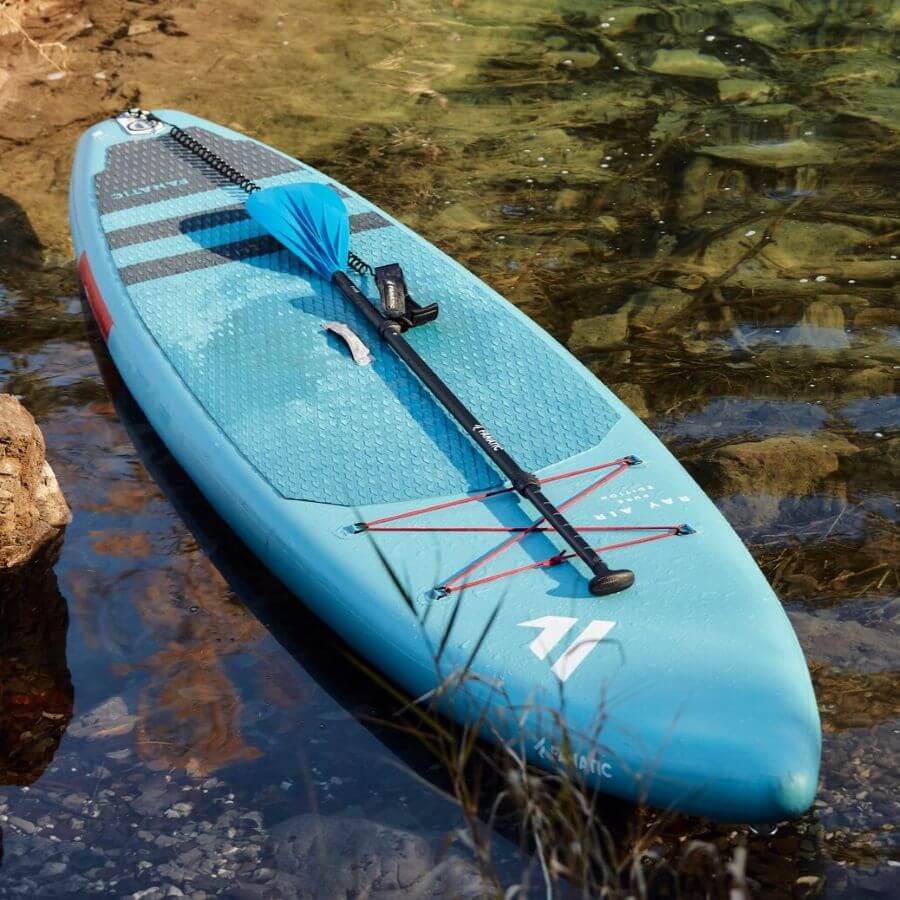 Blaues Stand-Up-Paddleboard mit Paddel auf klarem Wasser, umgeben von Felsen und Pflanzen.