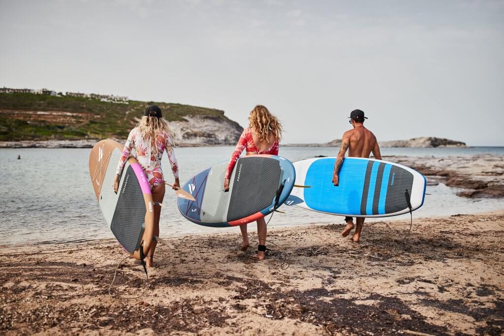 Drei Surfer, die Boards tragen, gehen an einem Strand entlang, mit ruhigem Wasser und Küstenlandschaft im Hintergrund.