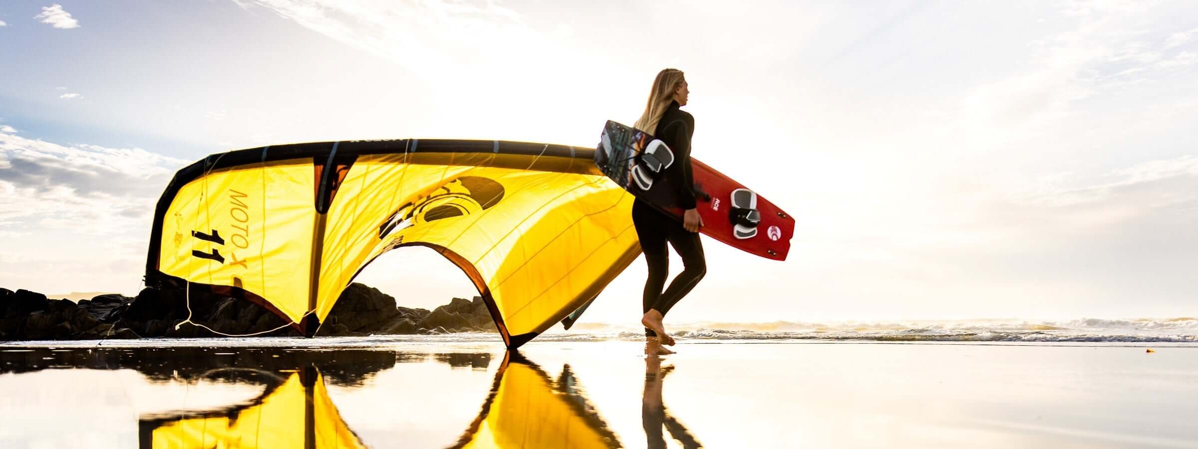 Surferin mit kitesurfing Ausrüstung läuft am Strand entlang, gelbes Kite-Segel reflektiert im Wasser.