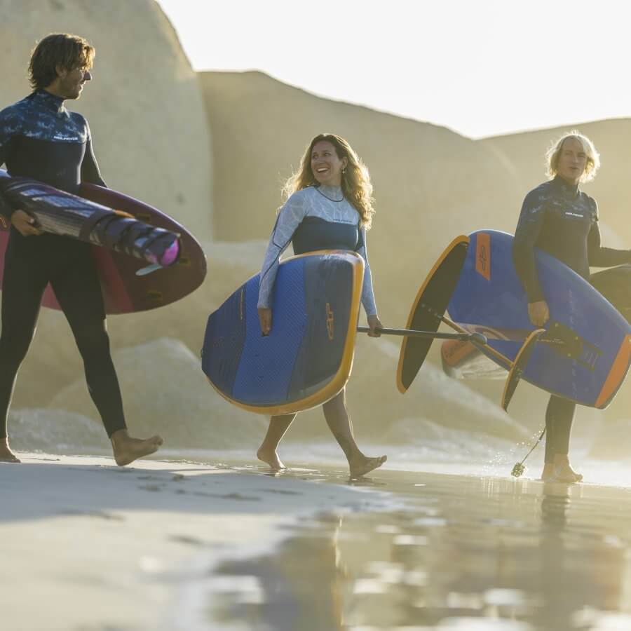 Drei Surfer tragen ihre Boards am Strand, strahlende Sonne und Wellen im Hintergrund. Sie tragen funktionale moderne Wassersportbekleidung.