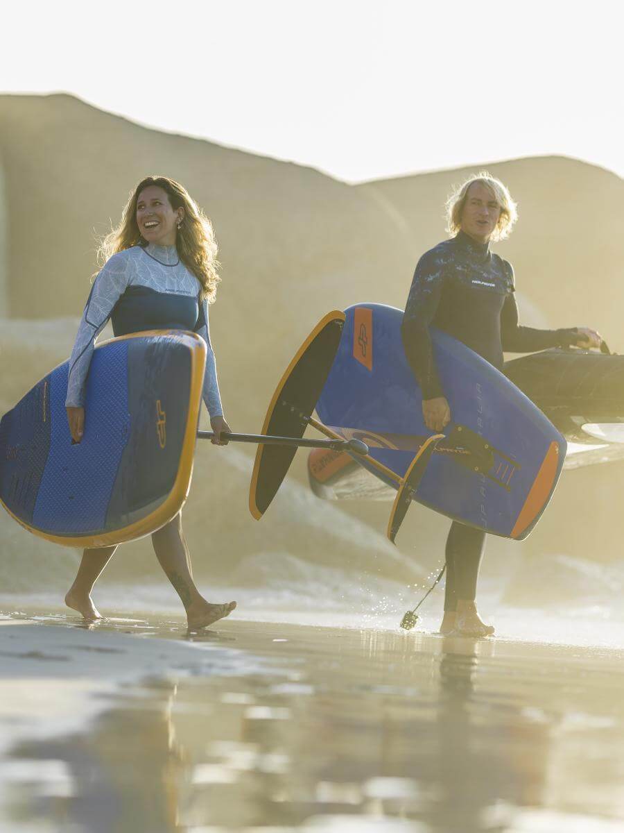Zwei Surfer tragen ihre Boards am Strand, strahlende Sonne und Wellen im Hintergrund.