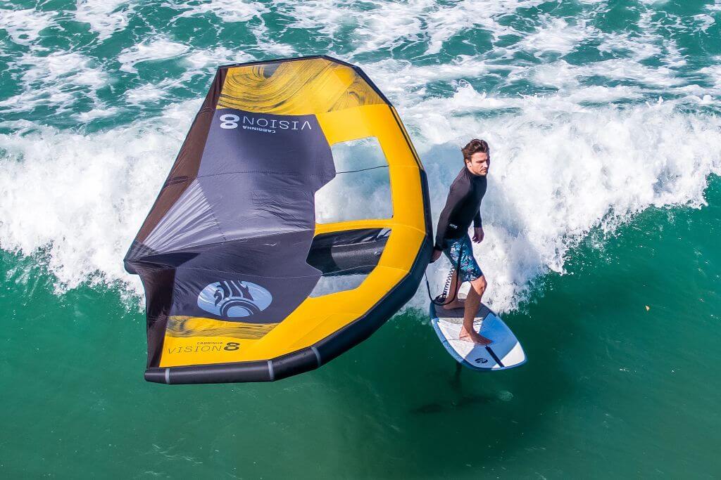 Surfer riding a wave with a wing sail on a sunny day, showcasing dynamic water sports action.